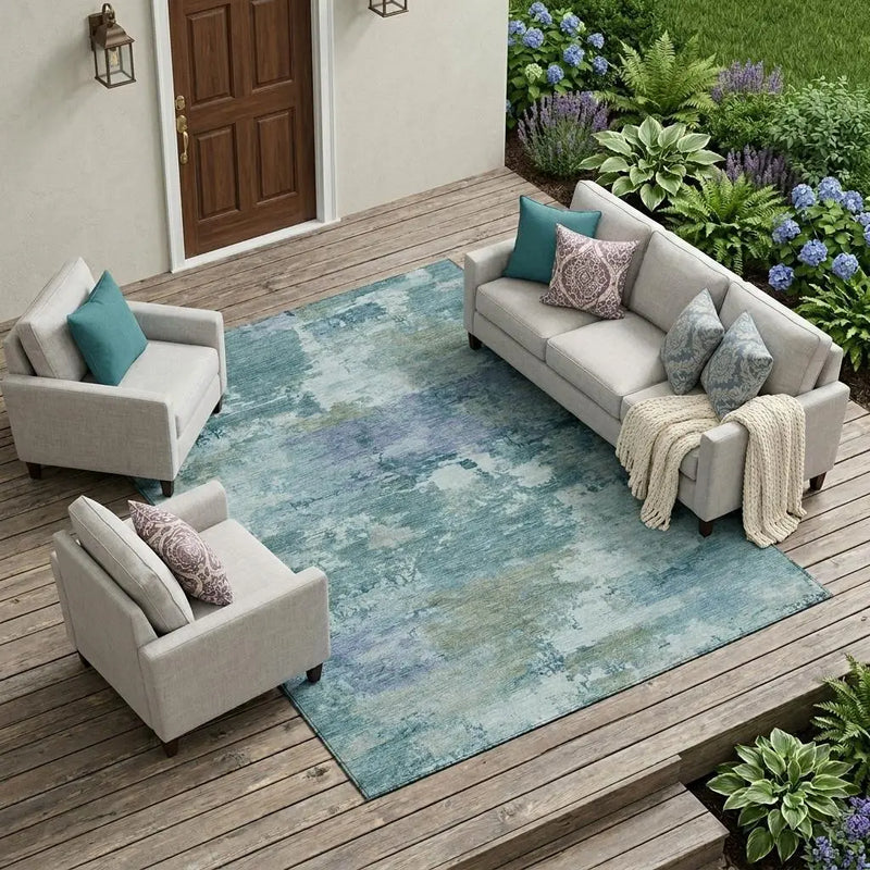 Light blue abstract area rug on a wooden patio with gray sofas and teal throw pillows, surrounded by lush greenery.