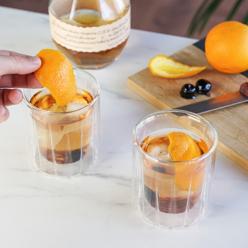 Orange peel garnish being placed into double-walled rocks glass with ice