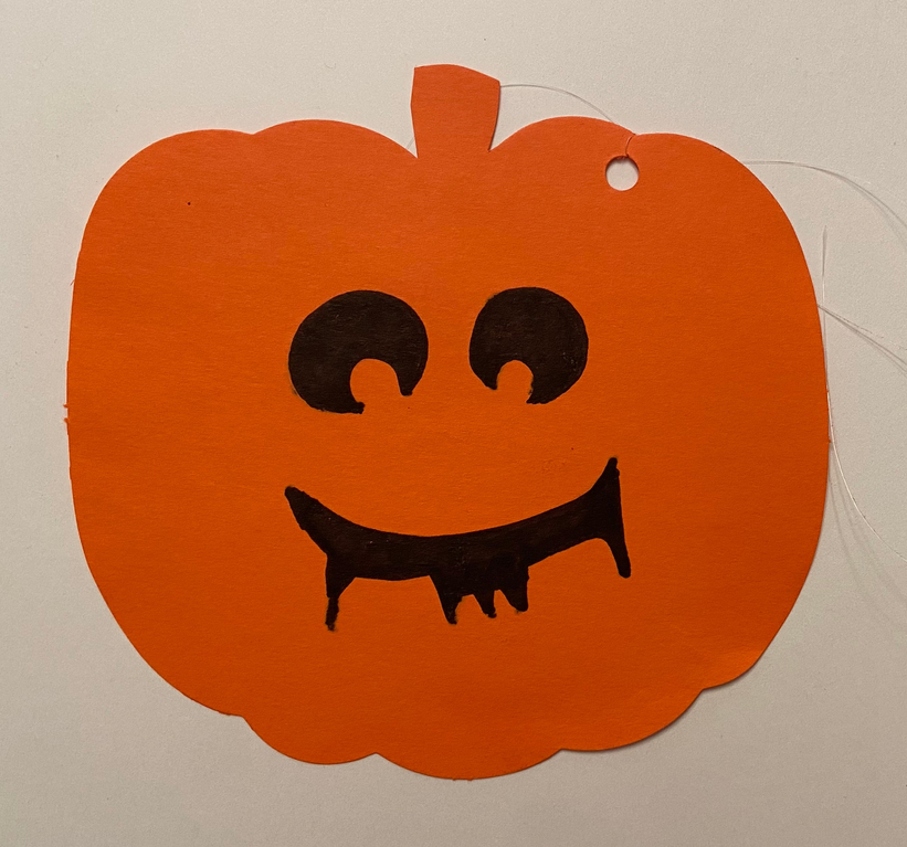 Orange jack-o’-lantern paper cut-out with black eyes, nose line, and smiling mouth