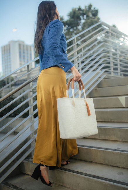 White woven tote bag with brown handles and tassel charm, carried on stairs