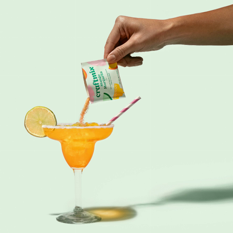 Hand pouring into orange cocktail glass with yellow mixer sachet