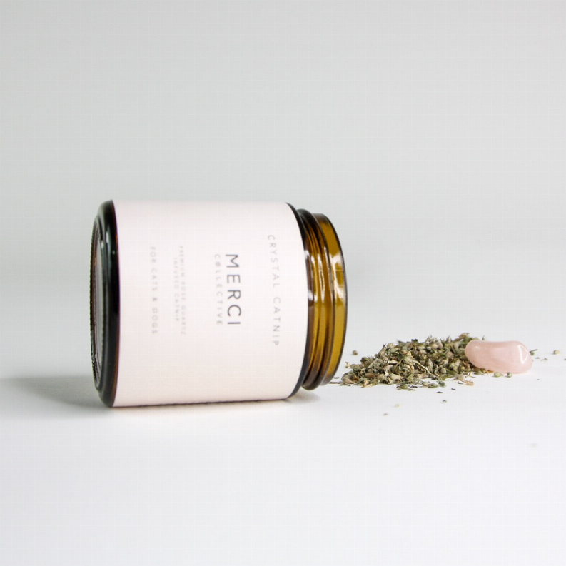 White catnip jar with black lid, gold rim; dried flakes and a pink crystal visible.