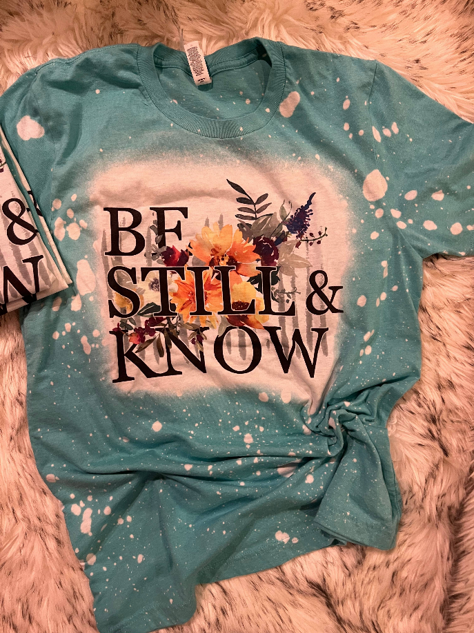 Dark teal crewneck T-shirt with large floral "Be Still & Know" graphic and paint splatter