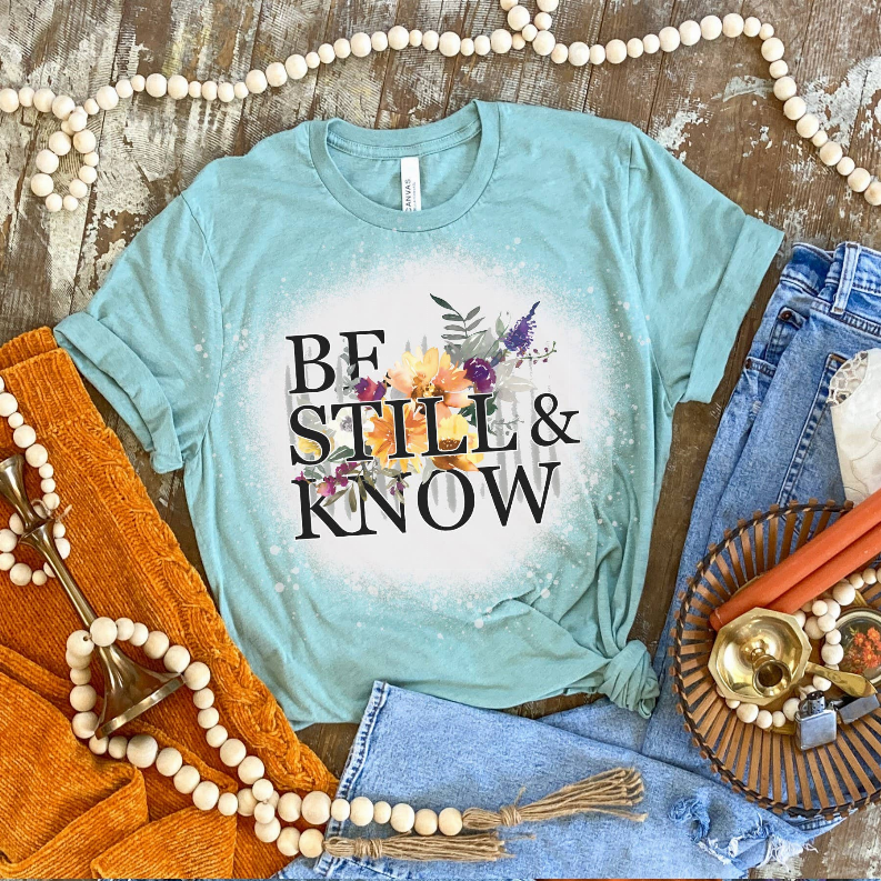 Teal crewneck graphic t-shirt with 'BE STILL & KNOW' text and flowers on the chest.