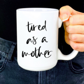 White ceramic coffee mug with black text "Tired as a mother" and curved handle