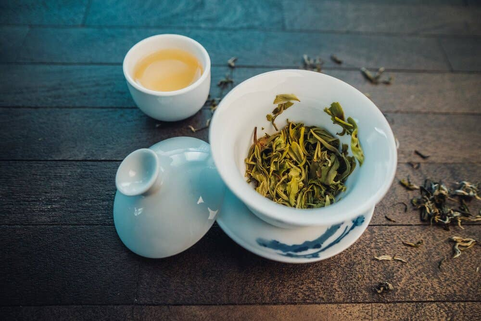 White bowl of loose green tea leaves with a small cup of brewed tea