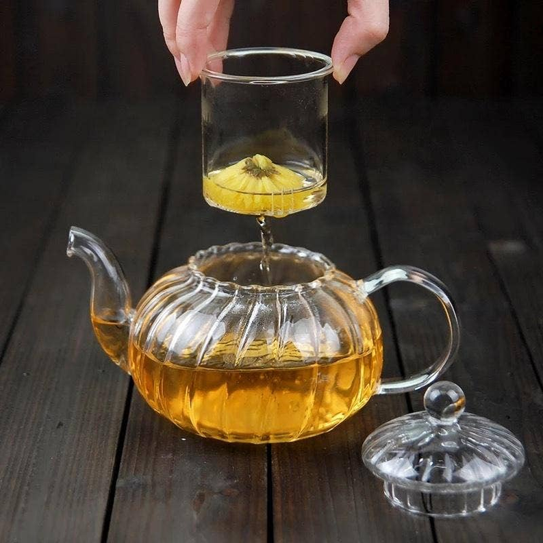 Clear glass teapot with lid and handles, holding amber tea and infuser basket