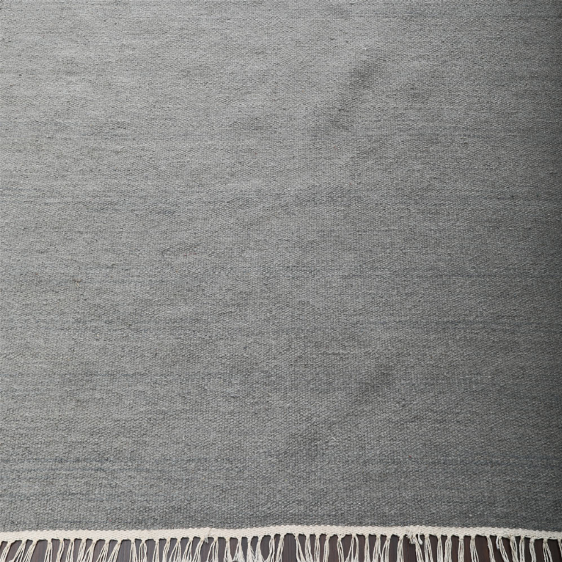 Gray woven rug surface texture with cream fringe trim along bottom edge
