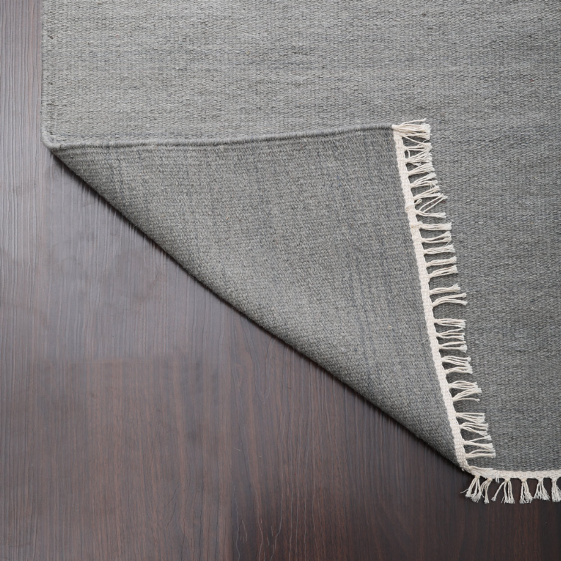 Gray flatweave wool rug corner with cream fringe and bound edge.