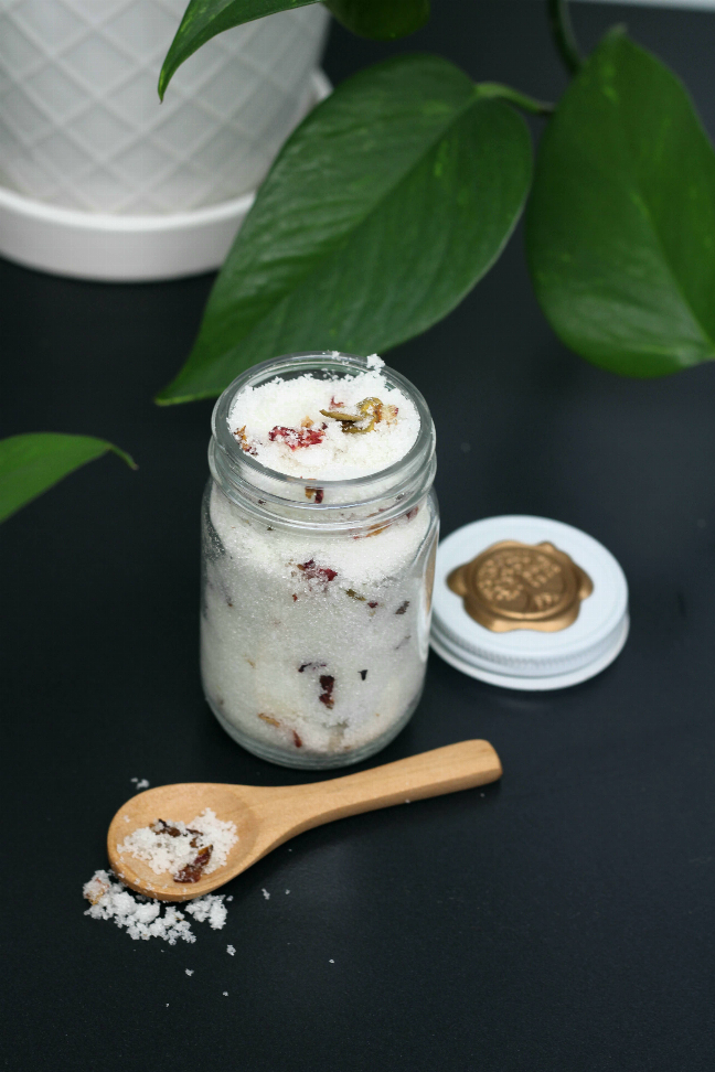 Open sugar scrub jar with wooden scoop and scattered sugar crystals
