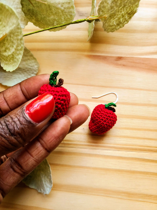 Red crocheted apple earrings with green leaves, held in hand