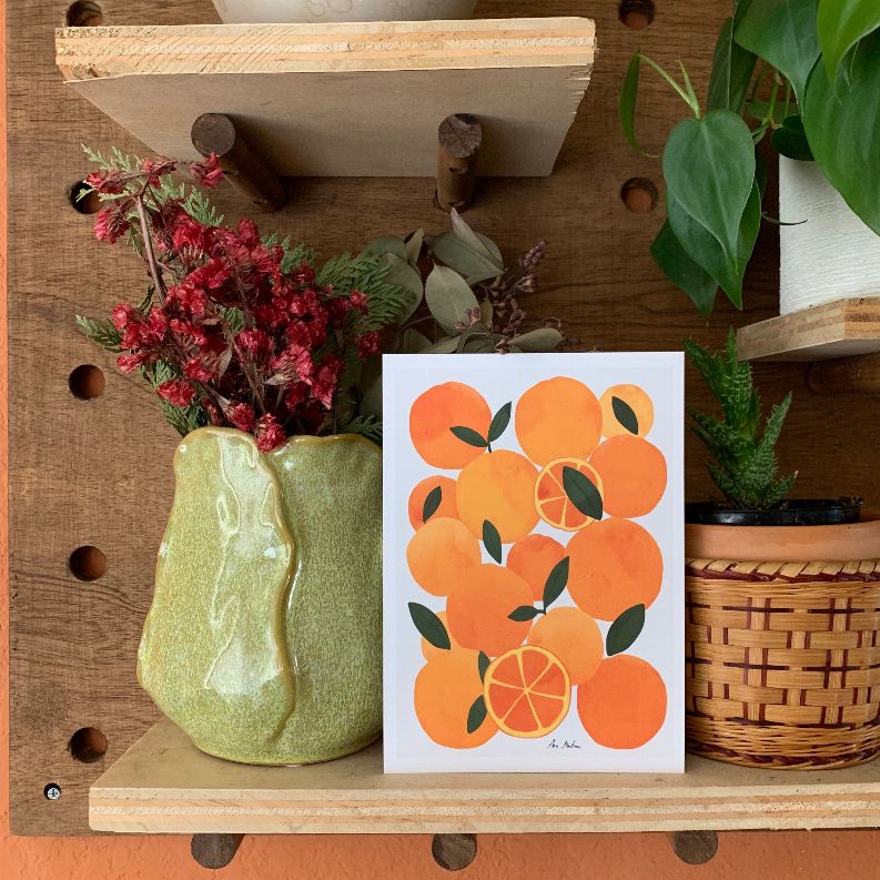 Orange fruit art print on a wooden shelf with plants and a woven basket