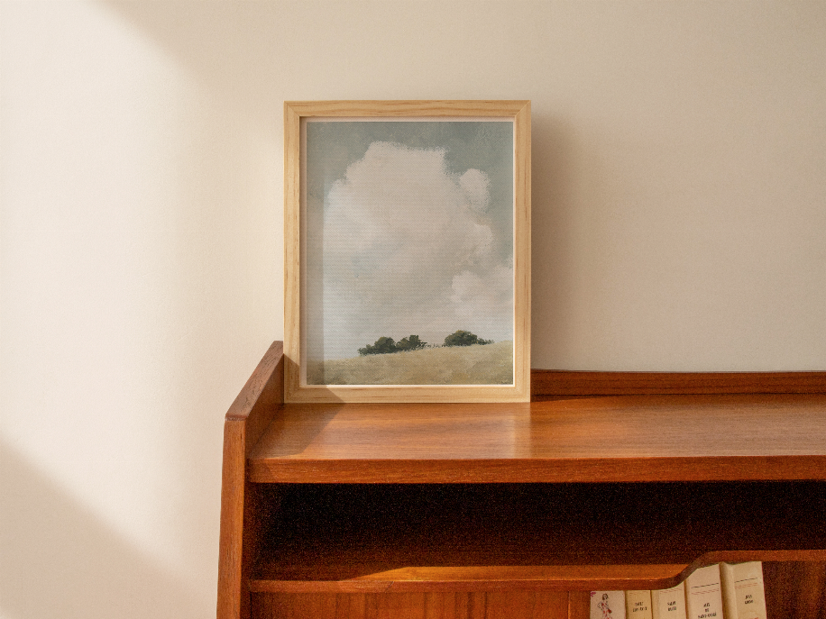 Wood framed landscape print on ledge, cloudy sky, green trees, and beige hill