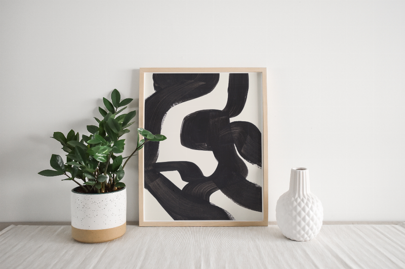 Framed black abstract ink art print with potted plant and white ceramic vase on table