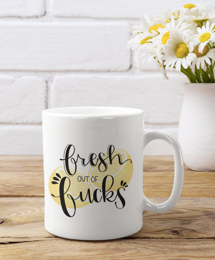 White coffee mug with 'Fresh out of Fucks' lettering and yellow heart graphic, angled view