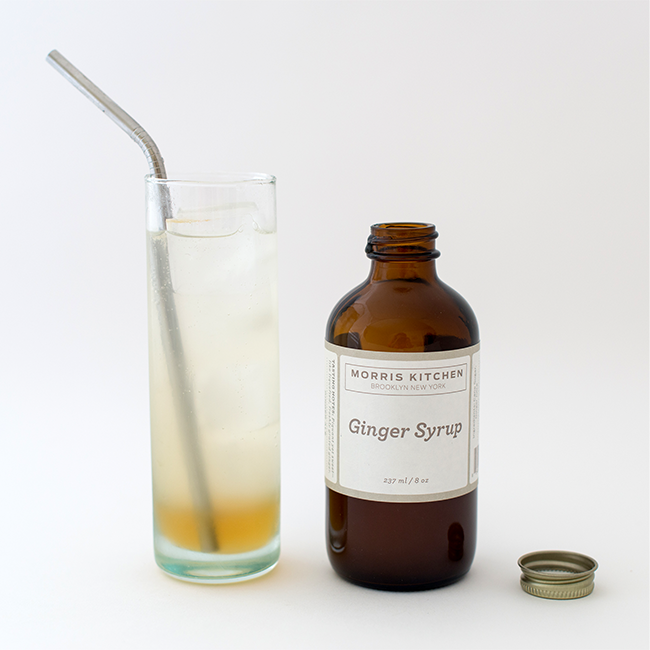 Ginger syrup bottle with gold cap beside tall glass with ice and straw