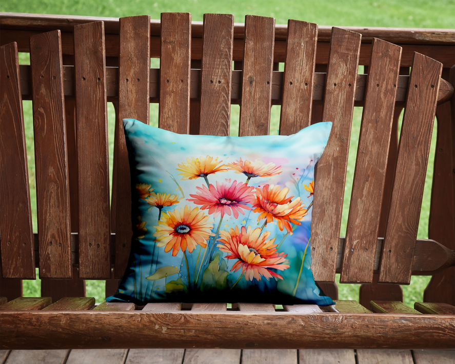 Watercolor floral throw pillow with bright gerbera-style flowers on a wooden outdoor bench.