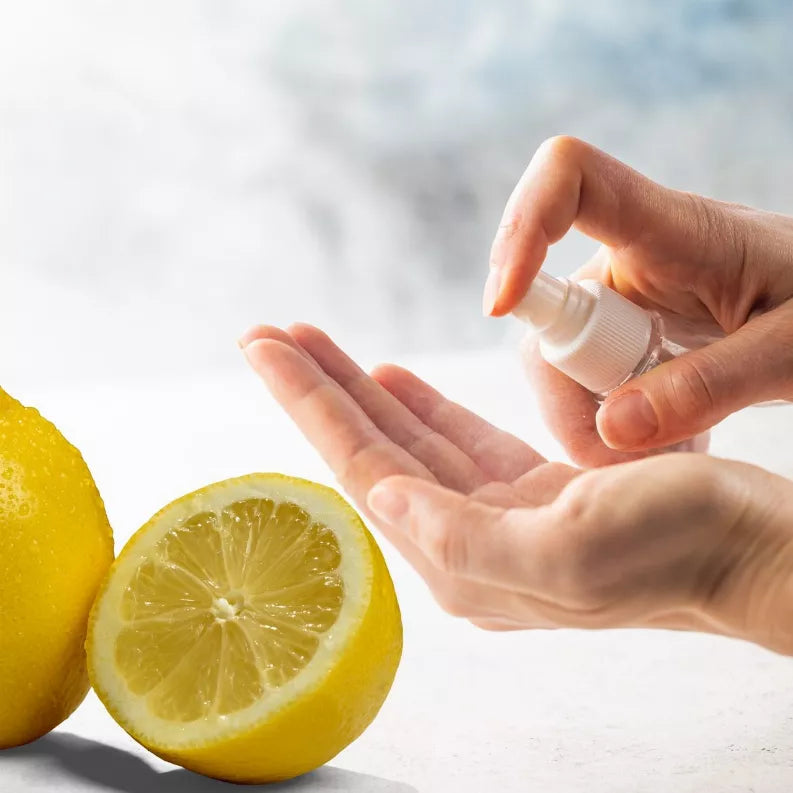 Hand holding travel-size sanitizing spray bottle above open palm beside lemon halves.