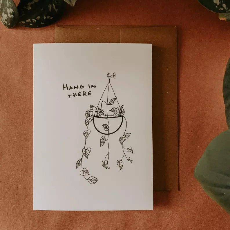 White greeting card with 'Hang in there' text and line-art hanging plant illustration.