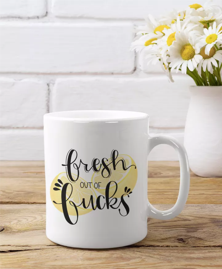 White ceramic coffee mug with 'Fresh out of Fucks' script and yellow emblem on front