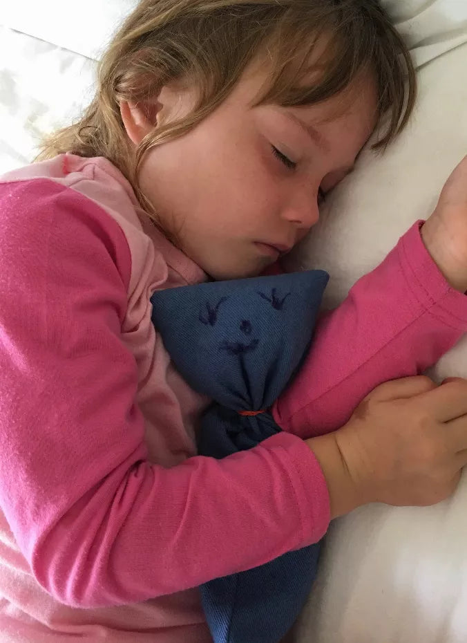 Blue fabric eye pillow between a child’s arms and face, rectangular cushion.
