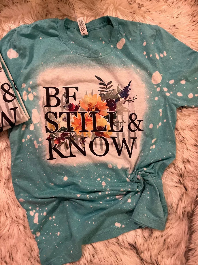 Dark teal short-sleeve graphic tee with “BE STILL & KNOW” floral speckle design