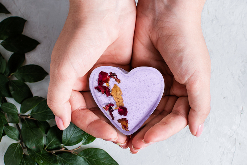 Hands holding a lavender heart-shaped shower steamer with red flower bits