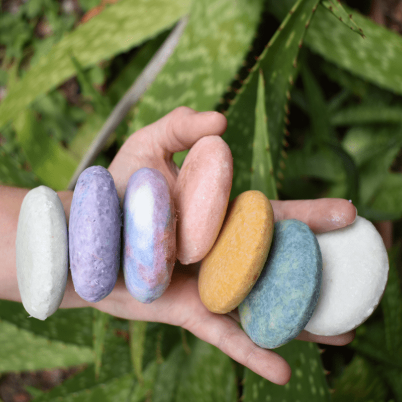 Hand holding assorted zero-waste shampoo bars in white, purple, peach, yellow, blue rounds