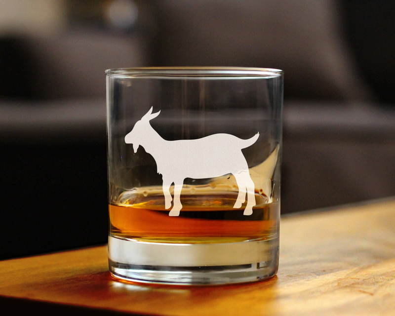 Whiskey rocks glass with hand-etched goat silhouette, standing on table with amber drink.