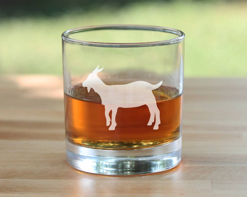 Whiskey rocks glass with hand-etched goat silhouette and amber drink, standing on table.