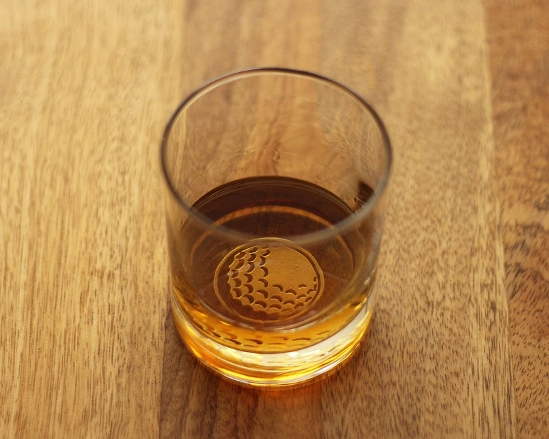Whiskey rocks glass, side view, with golf-ball emblem on base and amber liquid