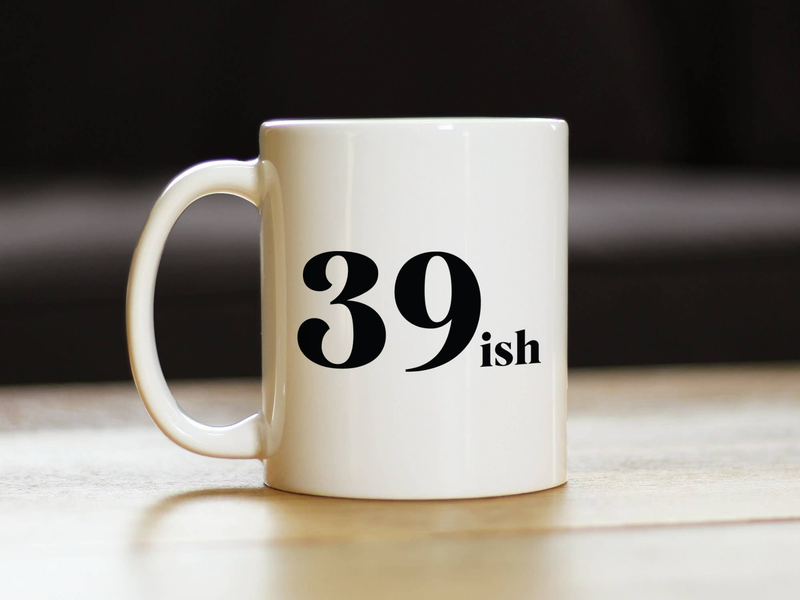 White ceramic coffee mug with black '39 ish' print, front view on wooden surface