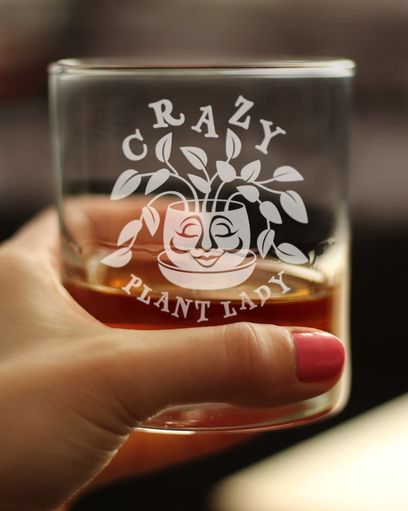 Whiskey rocks glass held in hand with etched Crazy Plant Lady logo; amber liquid.