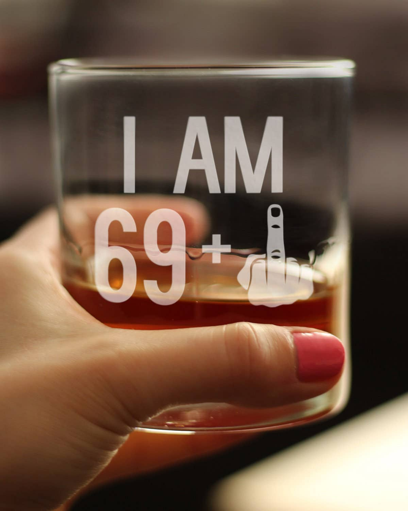 I AM 69+ middle finger whiskey rocks glass held in hand with amber liquid visible.
