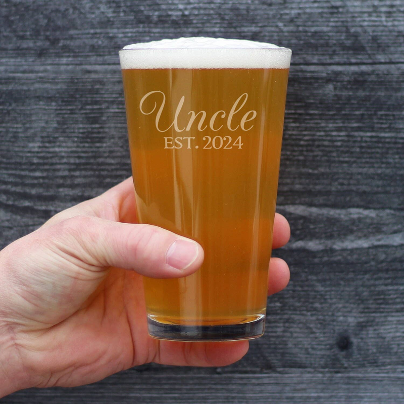 Hand holding Uncle EST. 2024 etched pint glass with amber beer and foam head