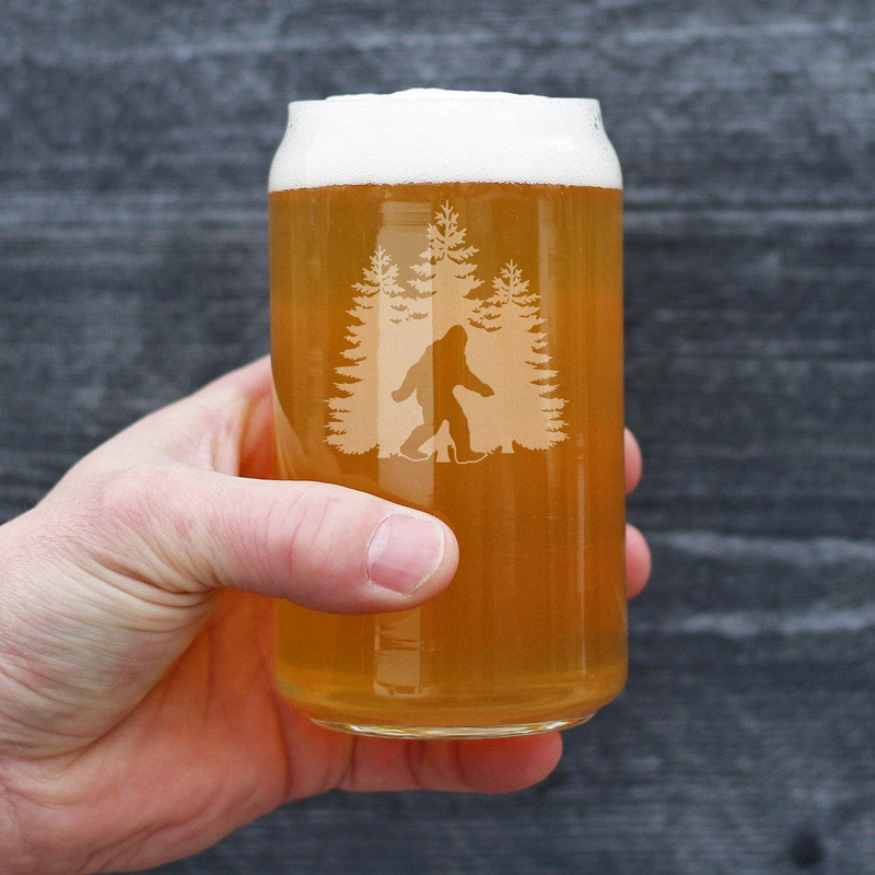 Hand holding pint beer can glass with amber beer and etched Sasquatch silhouette and pine trees