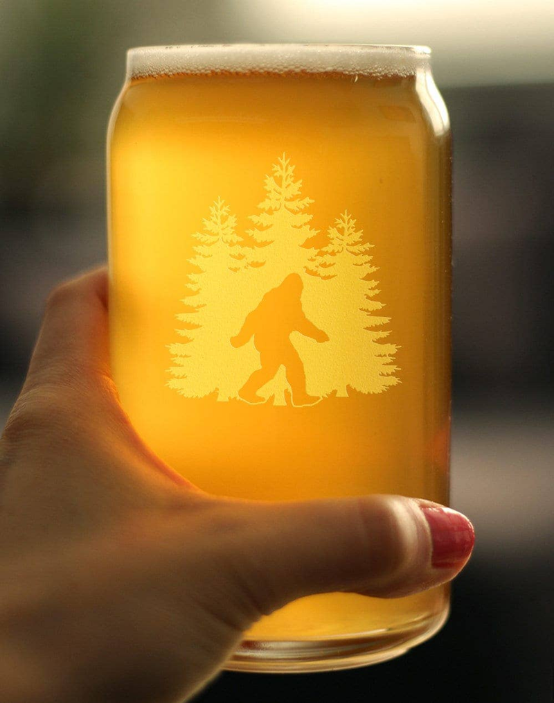 Hand holding pint beer can glass with amber beer and etched Sasquatch among pine trees