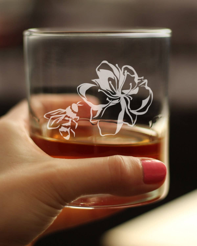 Hand holding a clear whiskey rocks glass with etched flower and bee design, amber drink