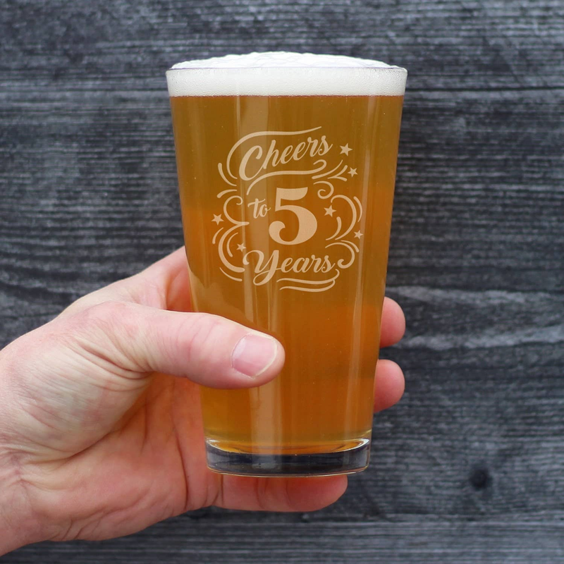 Hand holding clear pint glass with amber beer and Cheers to 5 Years etched design