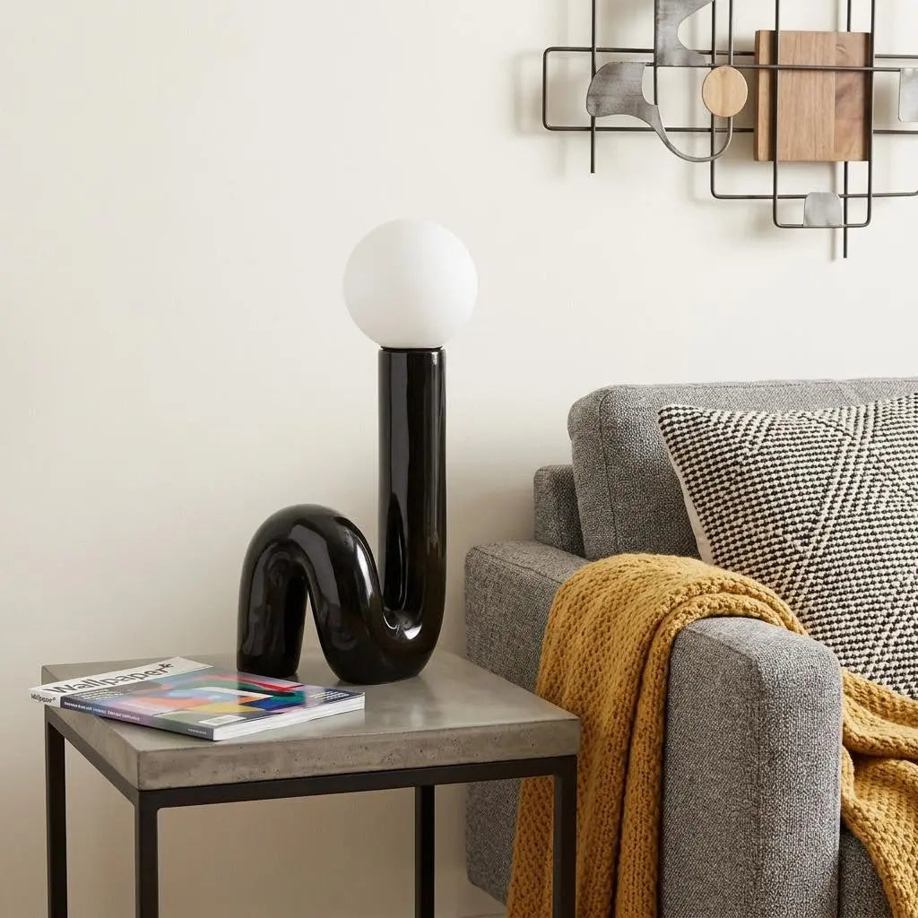 Modern black ceramic lamp with white globe, stylish curve design, beside a gray sofa and patterned throw blanket.