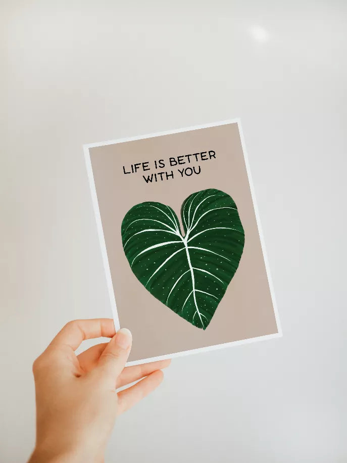 Handheld printed greeting card showing green leaf-heart and "LIFE IS BETTER WITH YOU"
