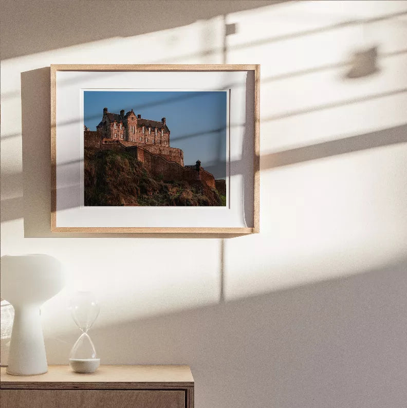 Framed castle print with light wood border, small fortress scene centered.