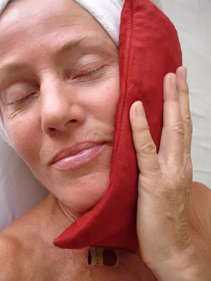 Red eye pillow held against one cheek, smooth rectangular cushion with side tag.