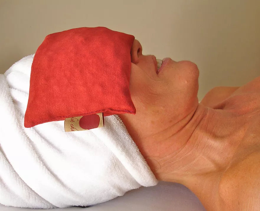 Red suede eye pillow resting on folded towel, rectangular cushion shape.