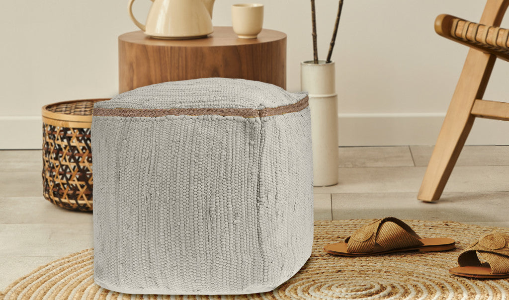 Light gray knitted pouf with a brown woven trim, placed on a natural fiber rug in a modern living space.