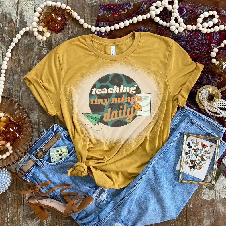 Mustard yellow crew-neck T-shirt with speckled splash effect behind “teaching tiny minds daily”