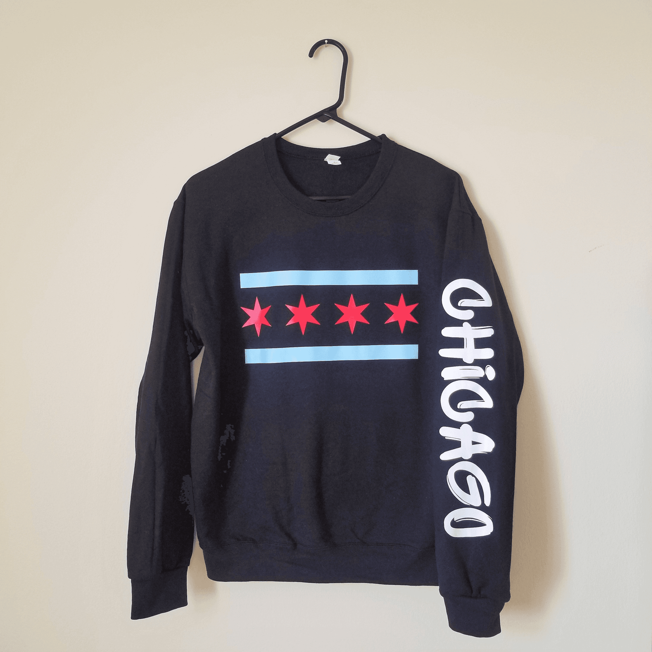 Dark crewneck sweatshirt on hanger with red stars, blue stripe, and white graffiti lettering