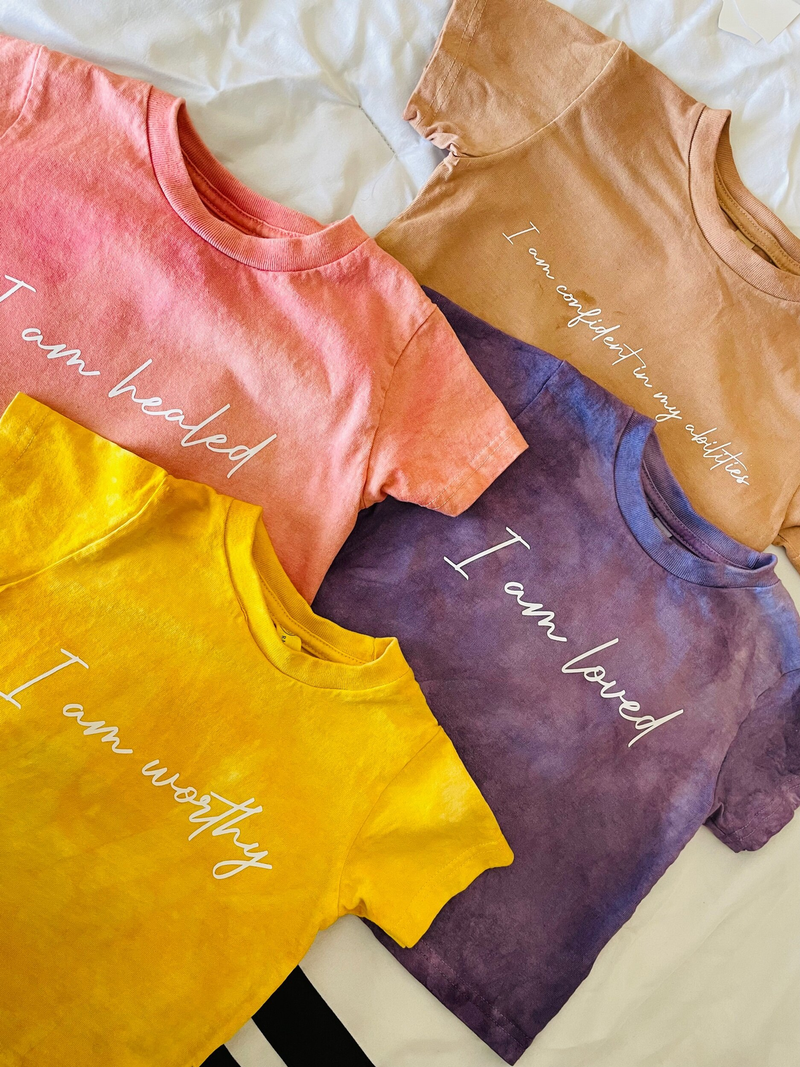 Flat-lay of four plant-dyed crewneck shirts: coral, yellow, purple, and tan with cursive print