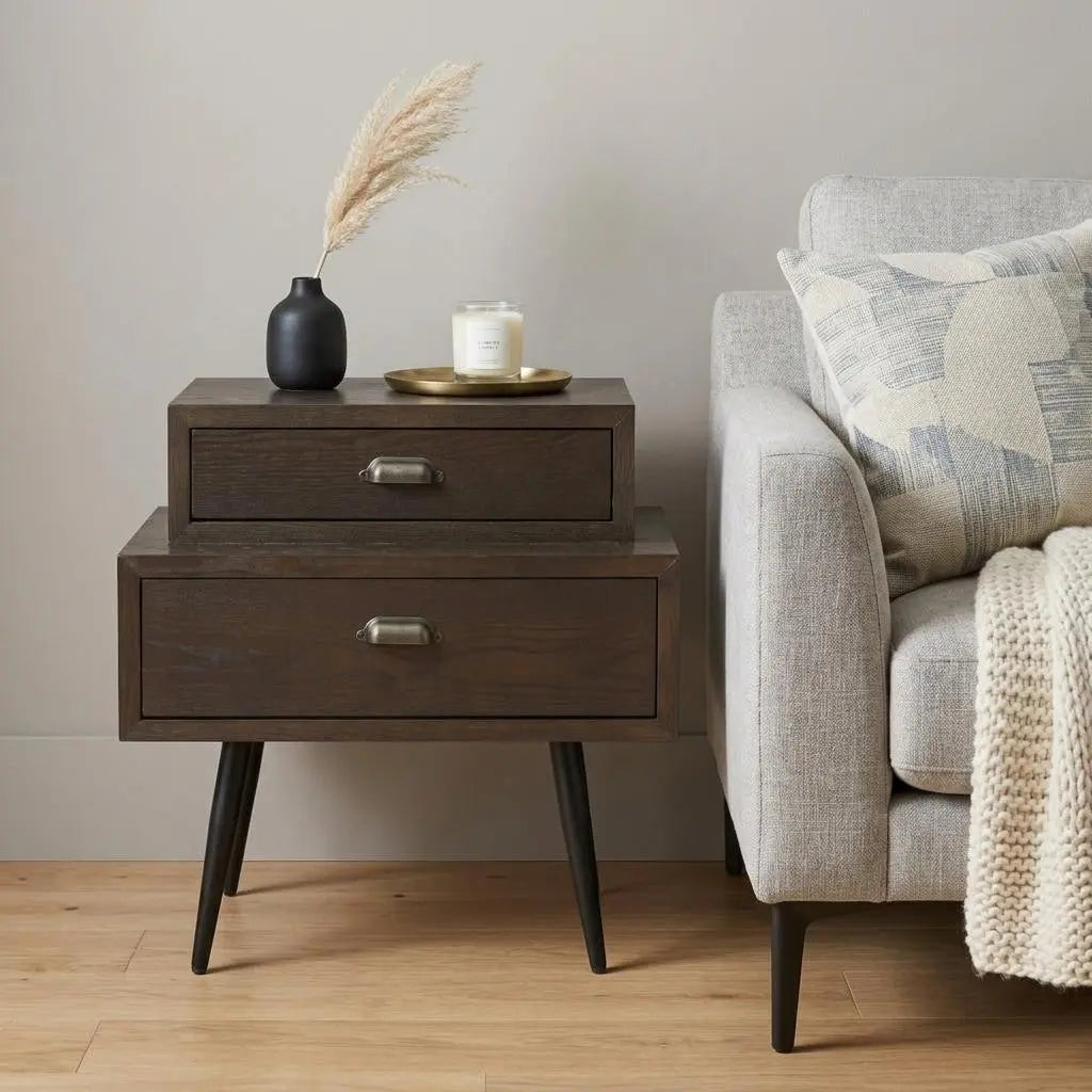 Dark wood nightstand with two drawers, black metal legs, styled with a vase and candle beside a gray sofa.