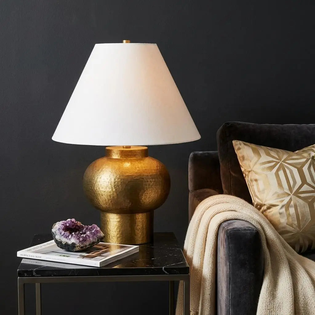 Gold hammered table lamp with white shade, modern design, placed on a black marble table beside a plush gray sofa.